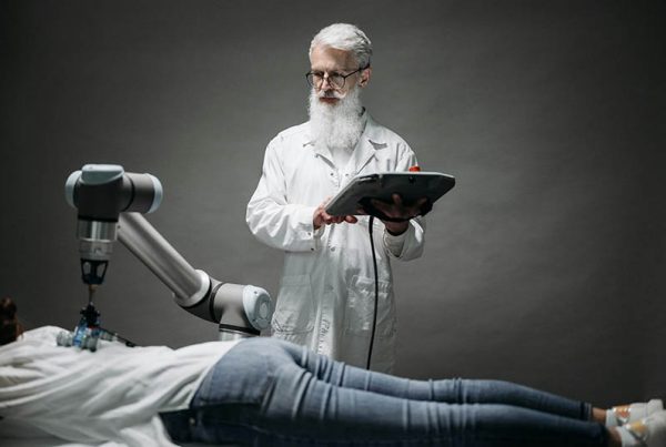 Doctor with beard, glasses and clipboard watches a robotic arm push into a patient's back